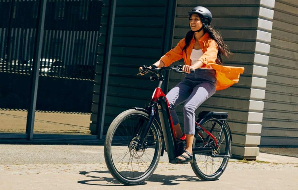 essoa andando de bicicleta elétrica em uma avenida arborizada, usando capacete, com sol da manhã refletindo no quadro da bike.